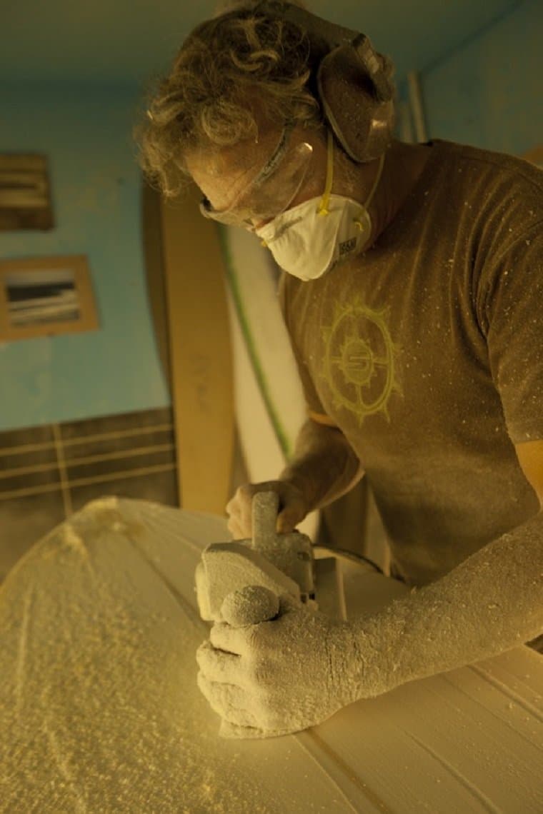 Tony Logosz planing a board blank in the shaping bay, goggles and dust mask on, foam dust on his arms