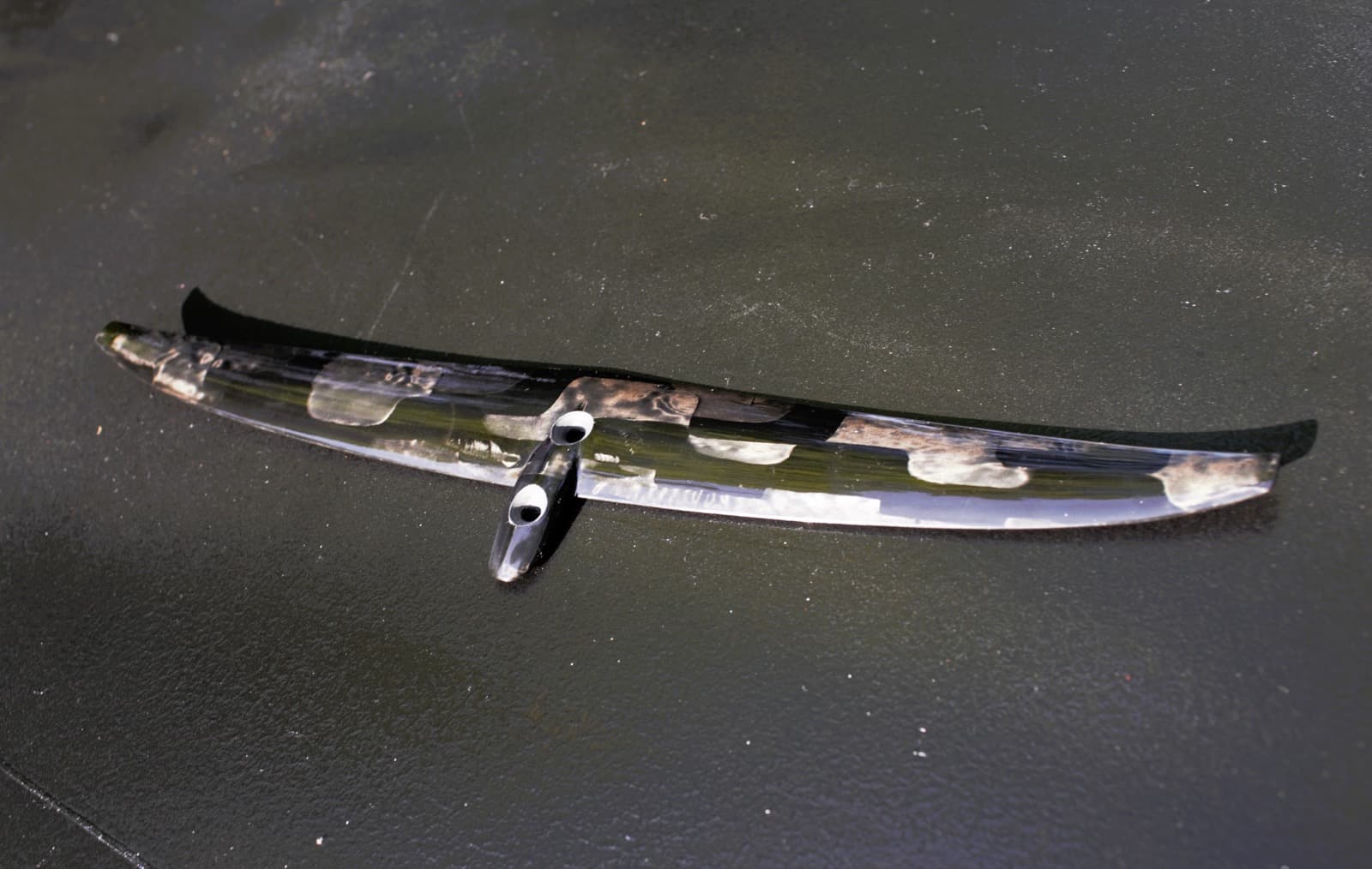 A carbon REVSTAR ST-A 140 hydrofoil stabilizer on a dark textured surface, seen from ahead and slightly above — a slim, nearly flat span with the carbon showing through a gloss finish, and the machined mounting pylon standing up at the centre with its two bolt holes facing the camera, which is the joint a stabilizer shim sits in